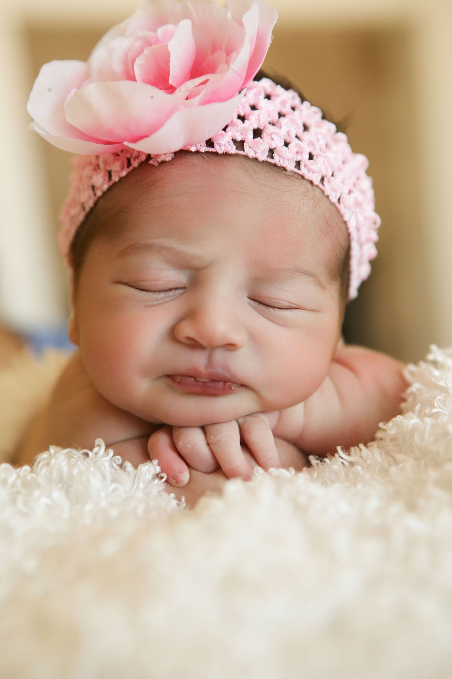 Sleeping newborn baby girl wearing a pink crocheted headband with a flower, resting chin on hands on a white fluffy blanket.