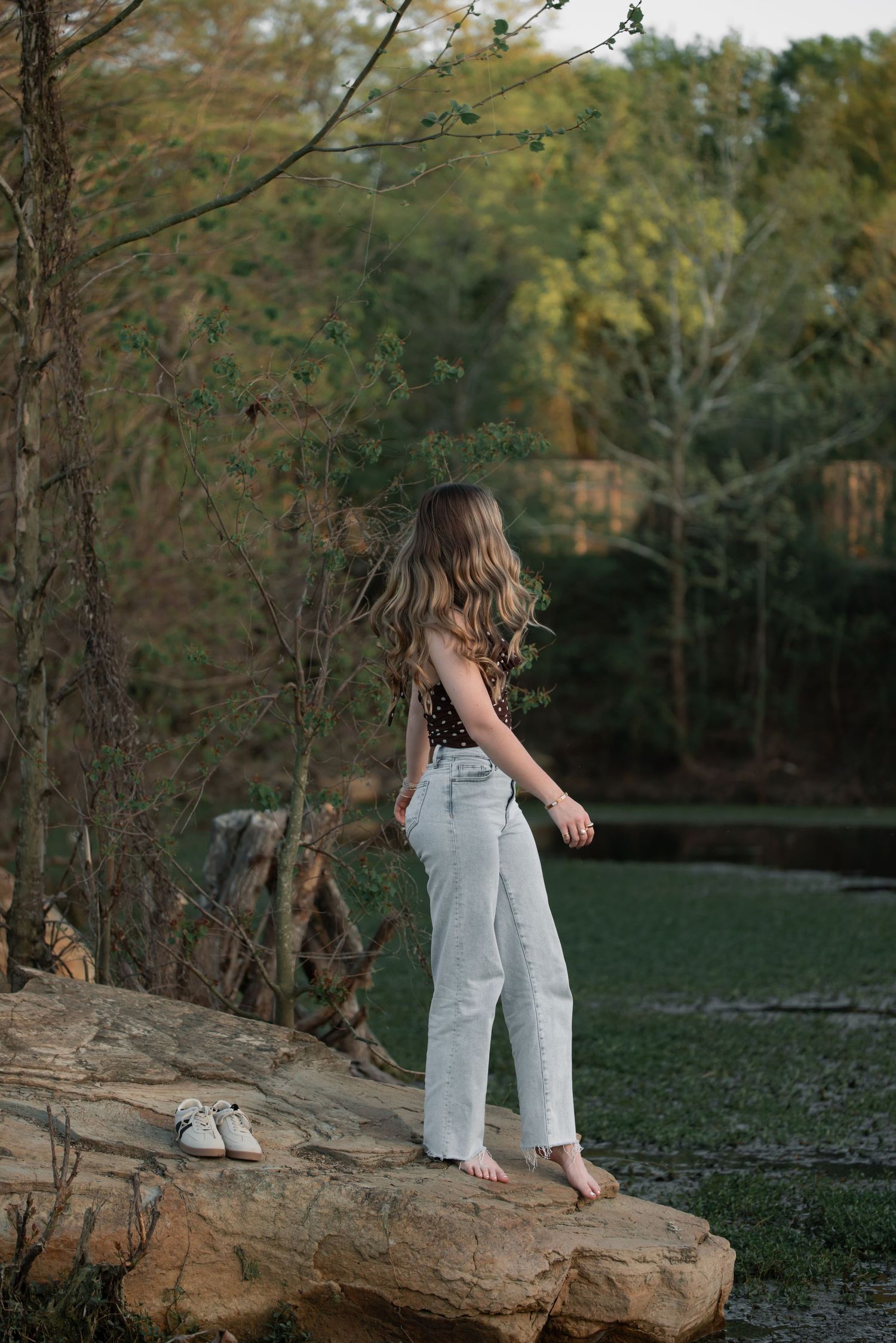 Young woman with long wavy hair stands barefoot on rocks in light jeans and dark top, surrounded by lush green trees.