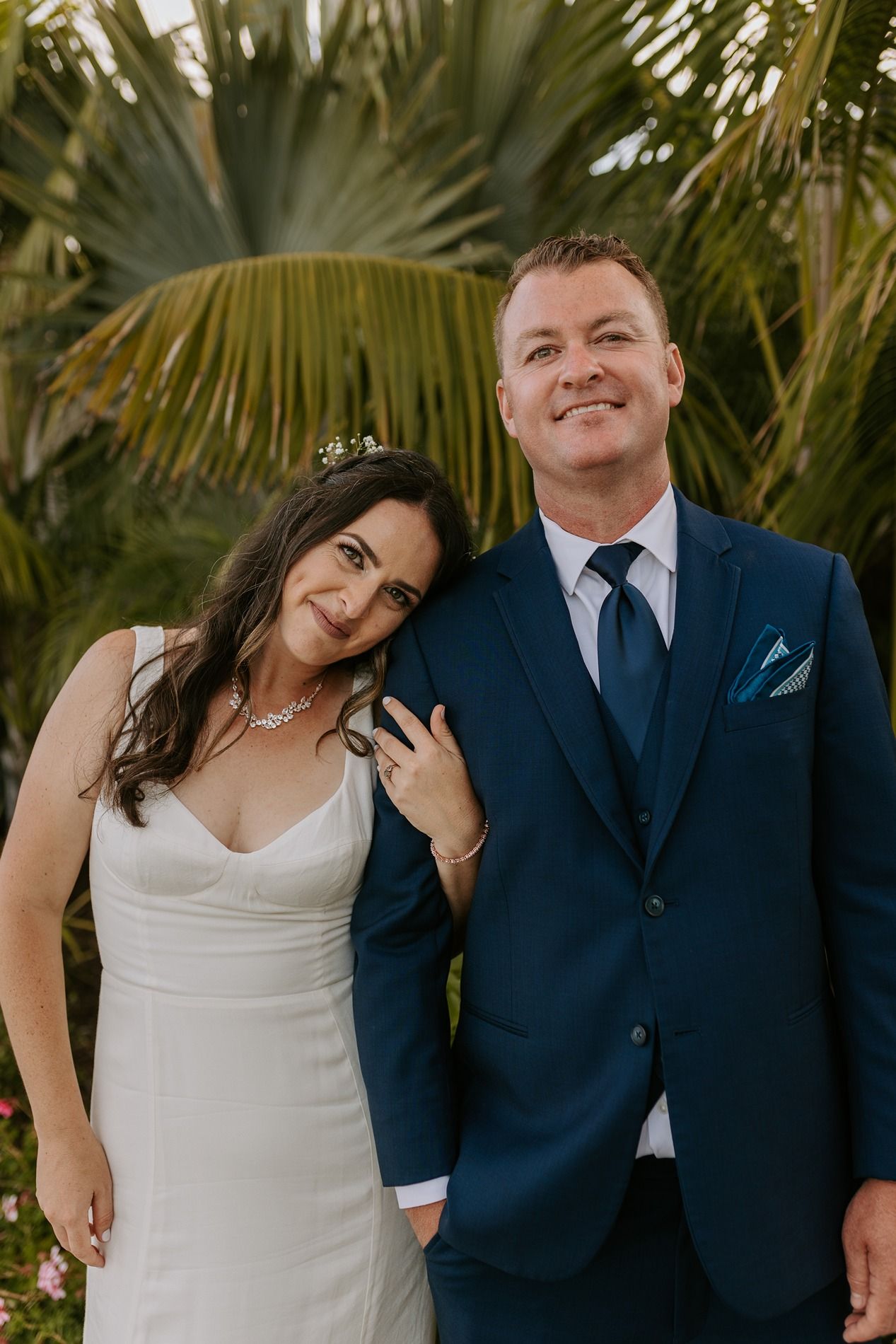 A bride in a white dress and groom in a navy suit smile together outdoors with tropical palm trees in the background at Tower Beach Club.