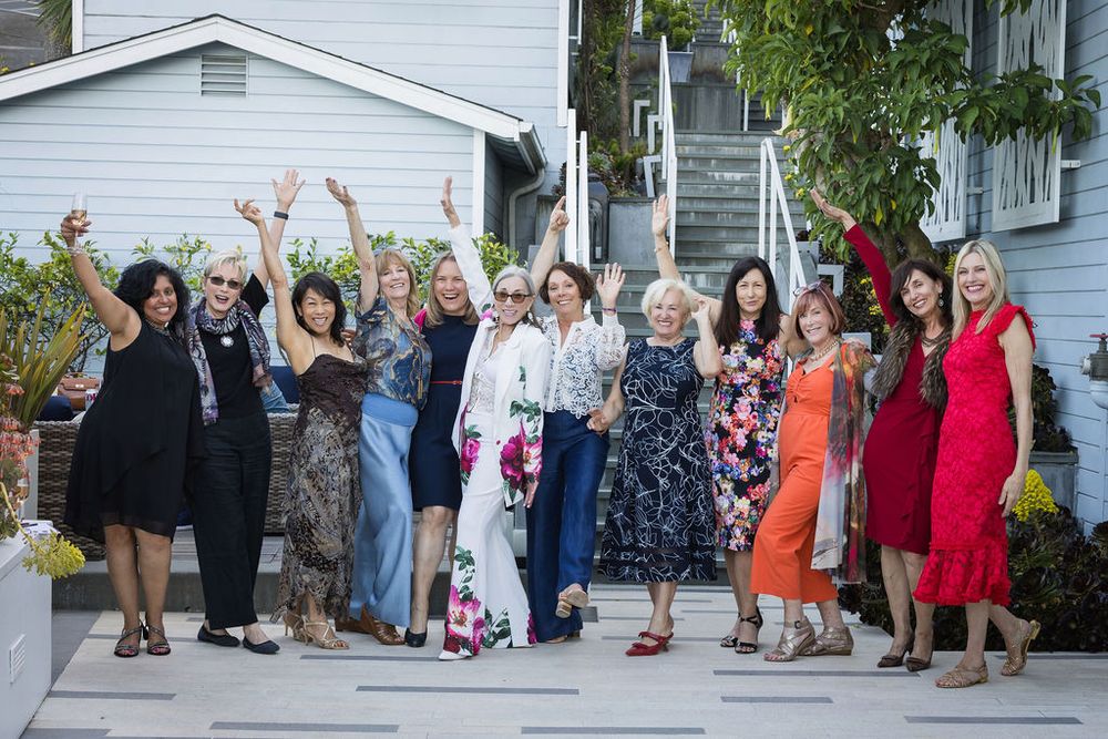 Group of women celebrating outdoors, raising hands joyfully in front of a white house.
