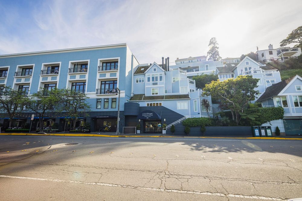 Blue multi-story hotel building on a quiet street with lush trees and hillside homes in the background on a sunny day.