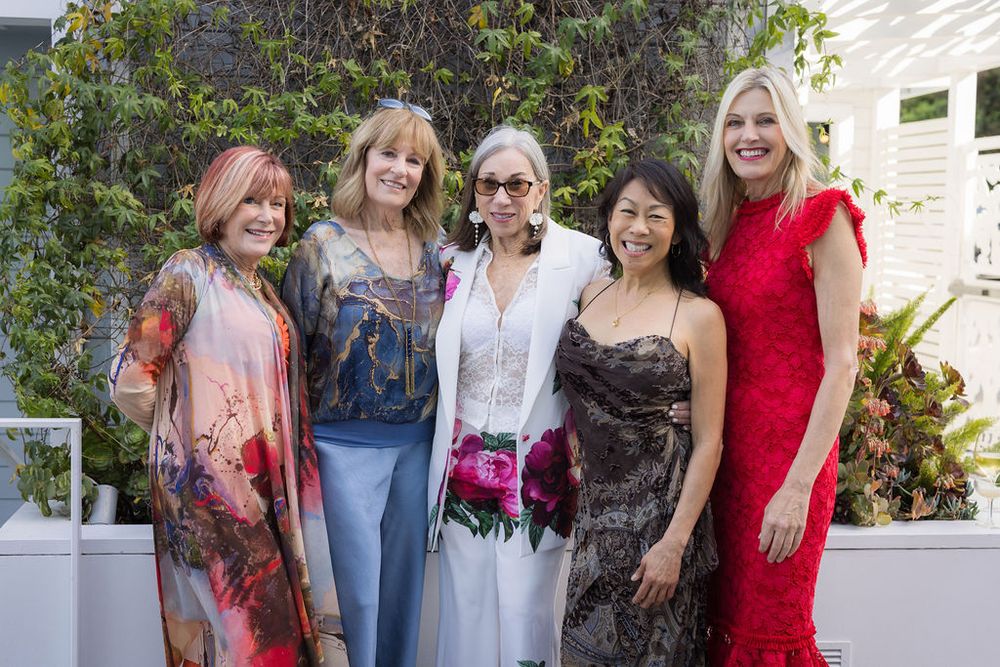 Five women smiling together outdoors at a garden event, dressed in elegant colorful attire.