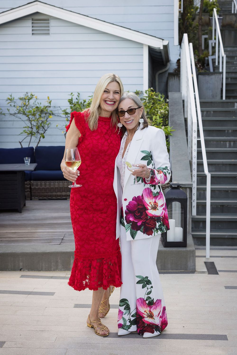 Two women in elegant attire holding wine glasses, posing outdoors at a social event.