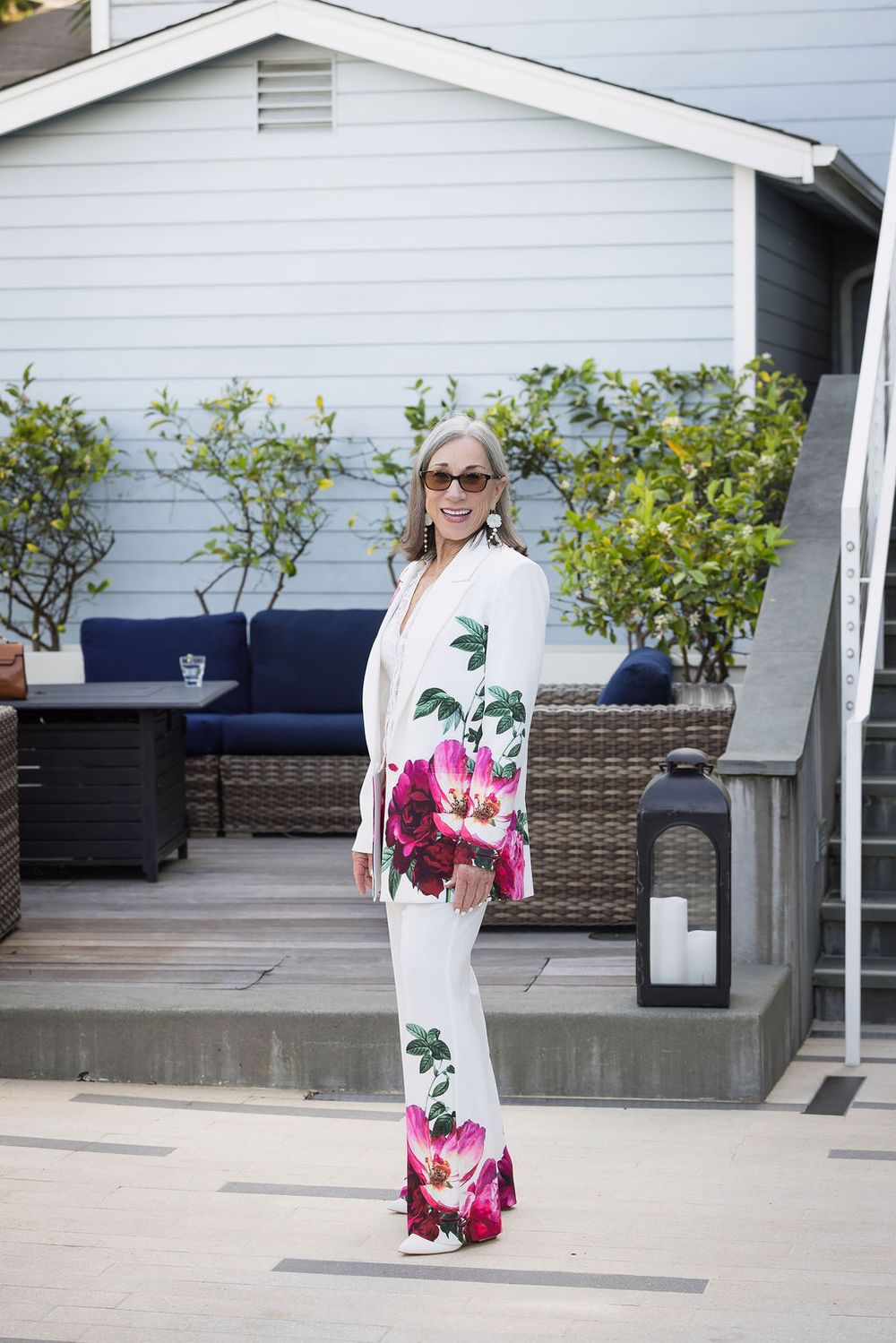 Woman in white floral jumpsuit posing on an outdoor patio with wicker furniture.