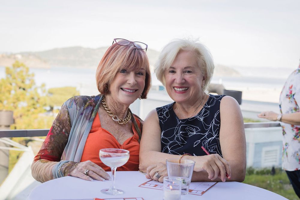 Two smiling women enjoy cocktails at an outdoor waterfront event, seated at a white table with scenic views.