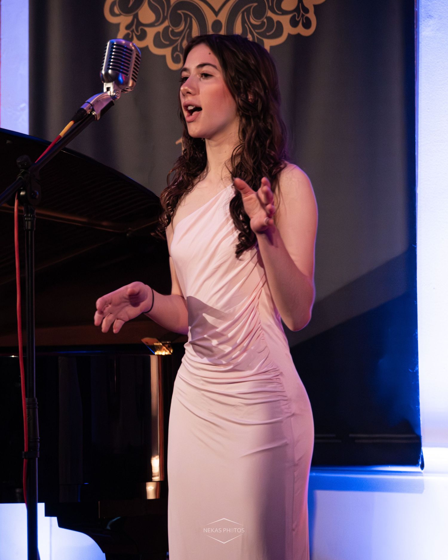 Young woman in white gown singing at vintage microphone on stage during elegant indoor performance.