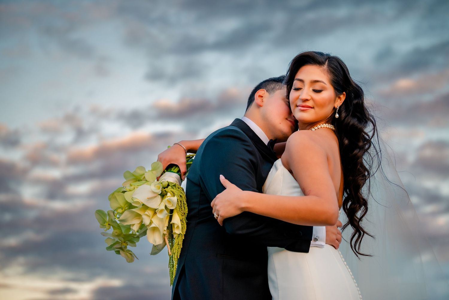 A romantic wedding couple embraces against a dramatic cloudy sunset sky while holding a green bouquet.