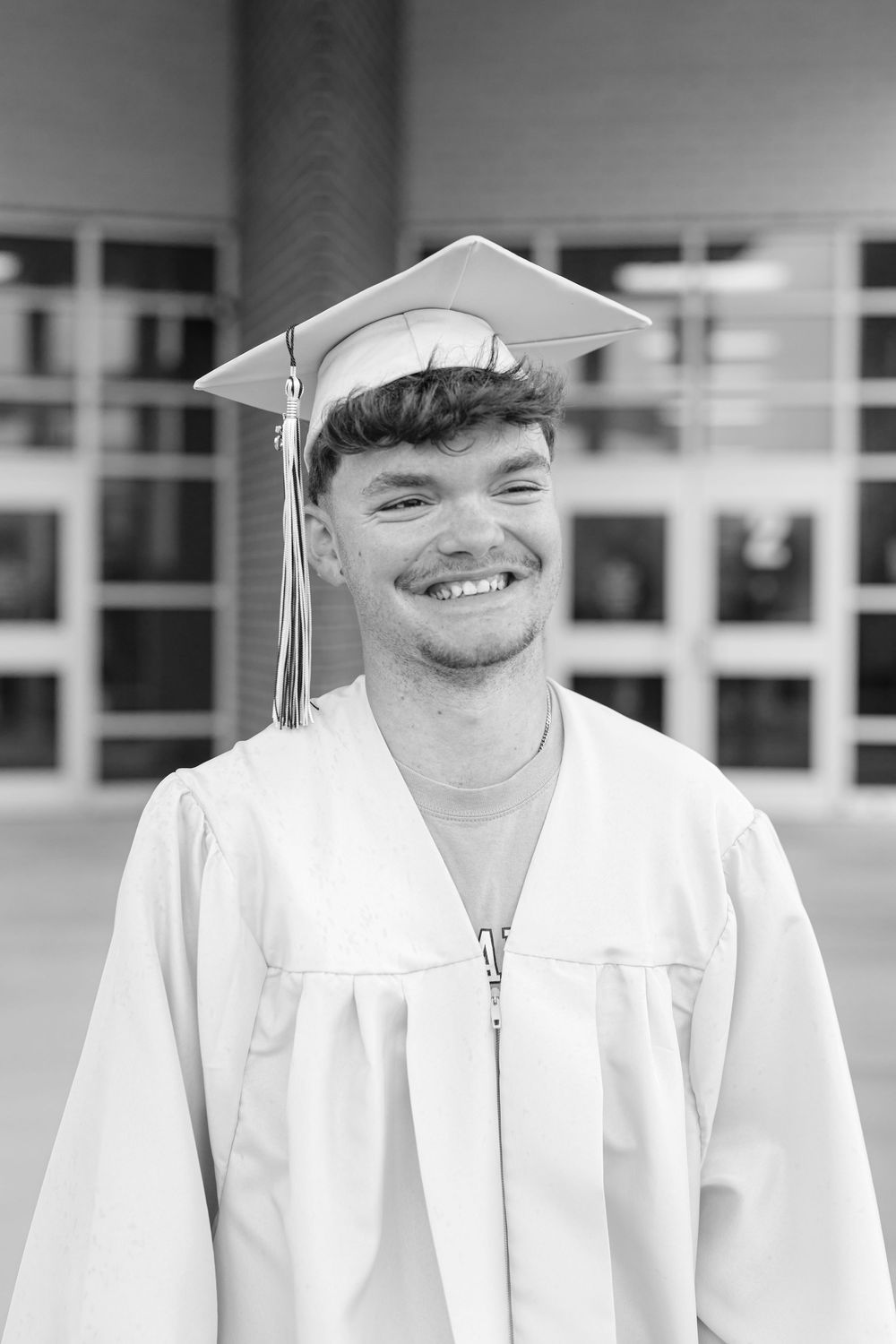 Smiling young man in white graduation cap and gown posing outside school building, black and white photo.