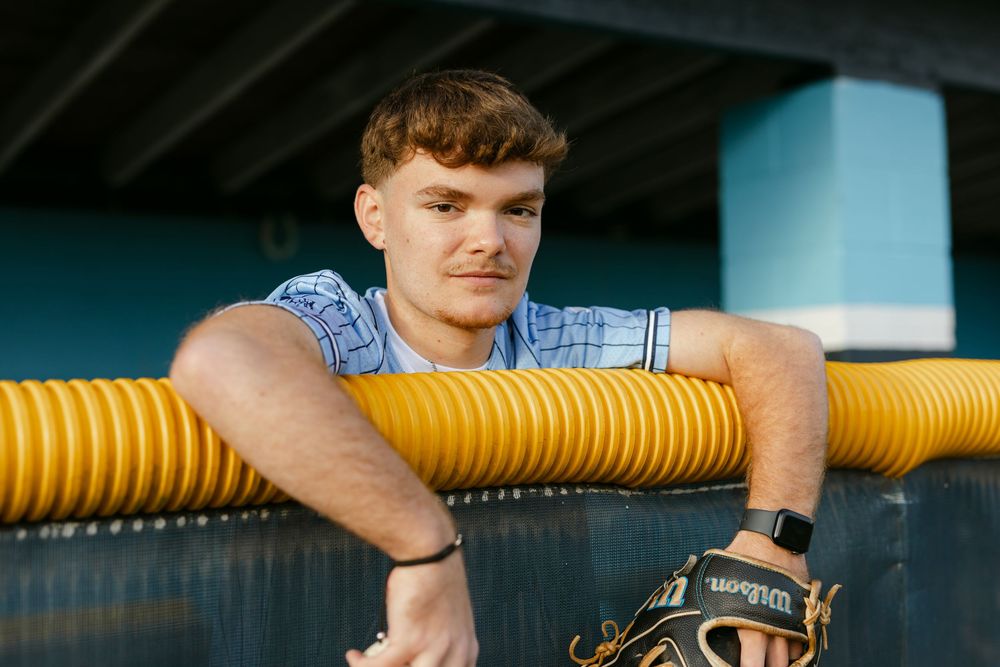 Young man in blue shirt leaning on yellow dugout railing holding Wilson baseball glove.