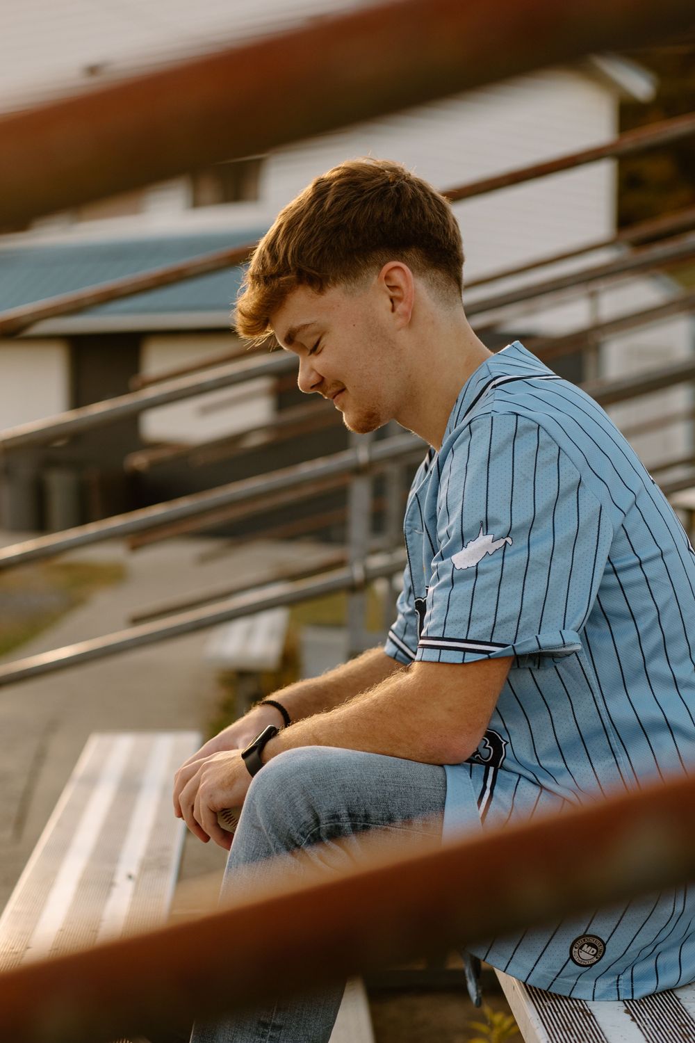Teen in blue pinstripe baseball jersey sitting on bleachers looking down in warm sunset light.