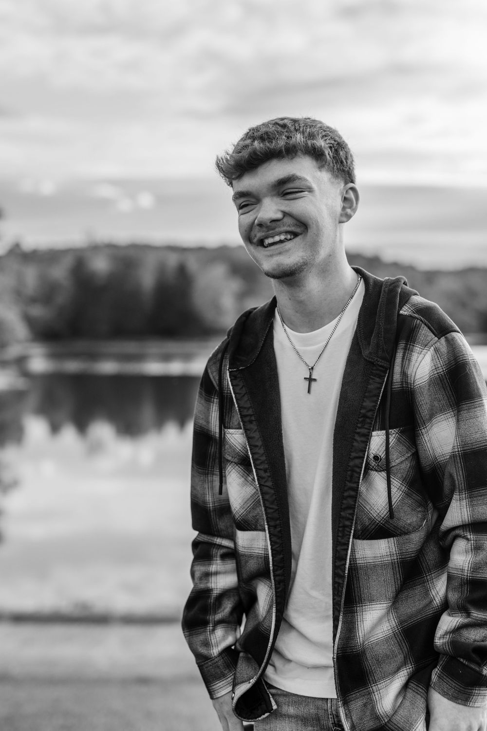 Smiling young man in plaid shirt and cross necklace laughing outdoors by a lake in black and white photo.