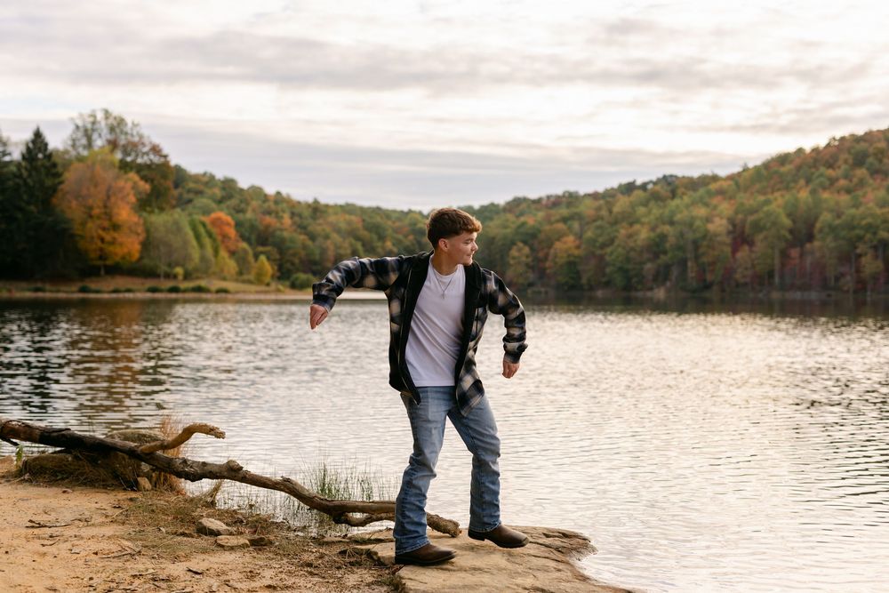 Young man in plaid shirt standing by a lake with vibrant fall foliage in the background.