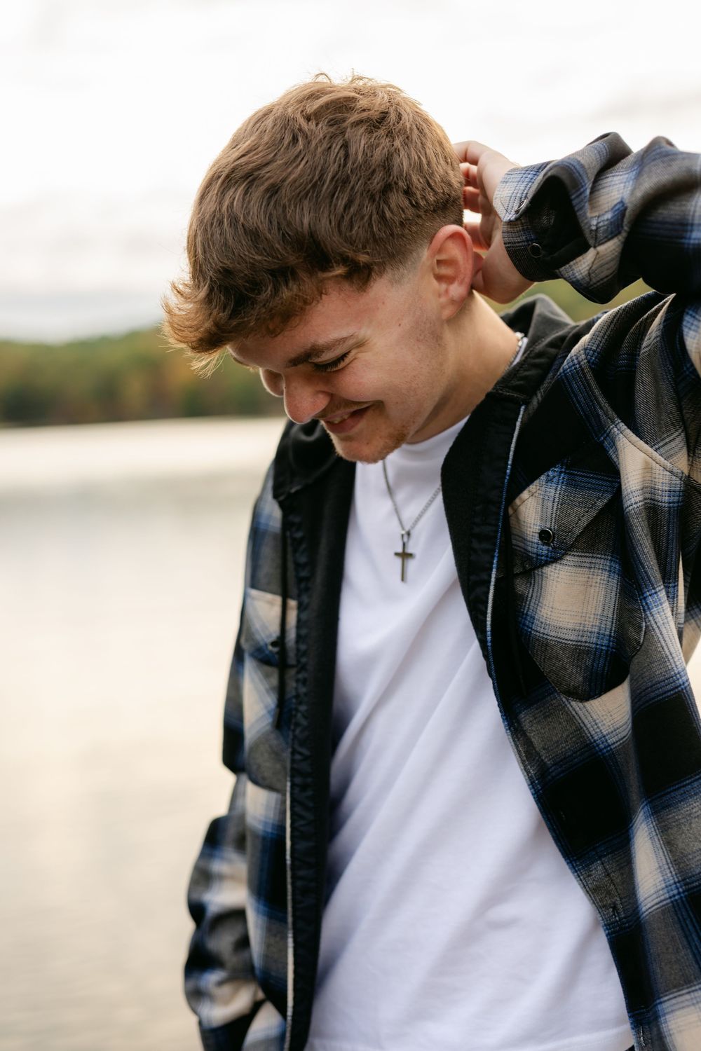 Laughing young man in plaid shirt and cross necklace standing near a lake on overcast day.