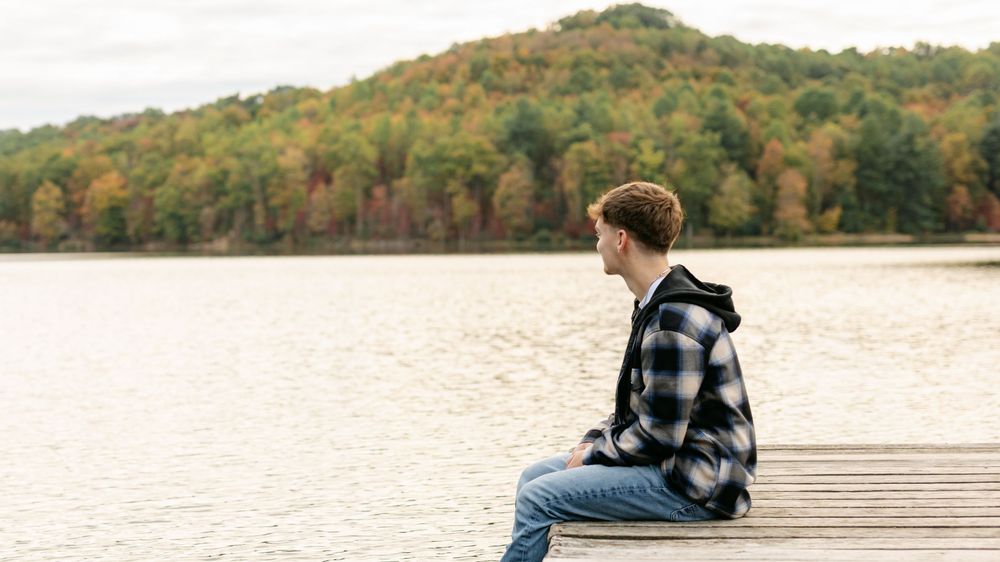 Teenage boy in plaid jacket sitting alone on a dock gazing at a lake with fall foliage.