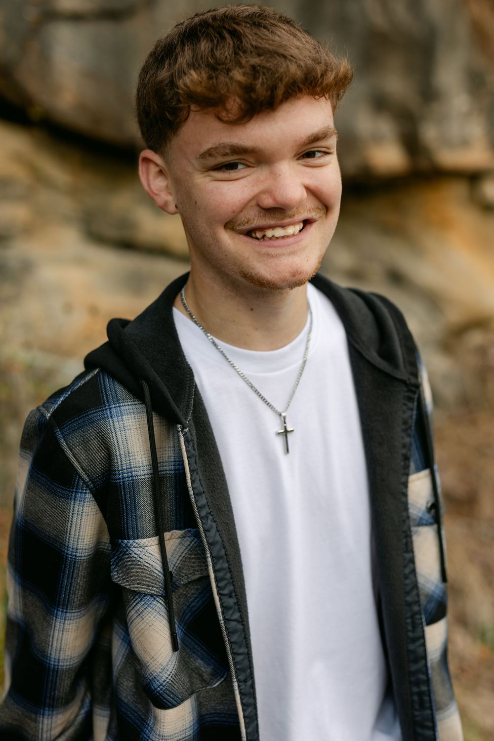 Smiling teenage boy wearing a cross necklace and plaid shirt posing in front of a rock wall.