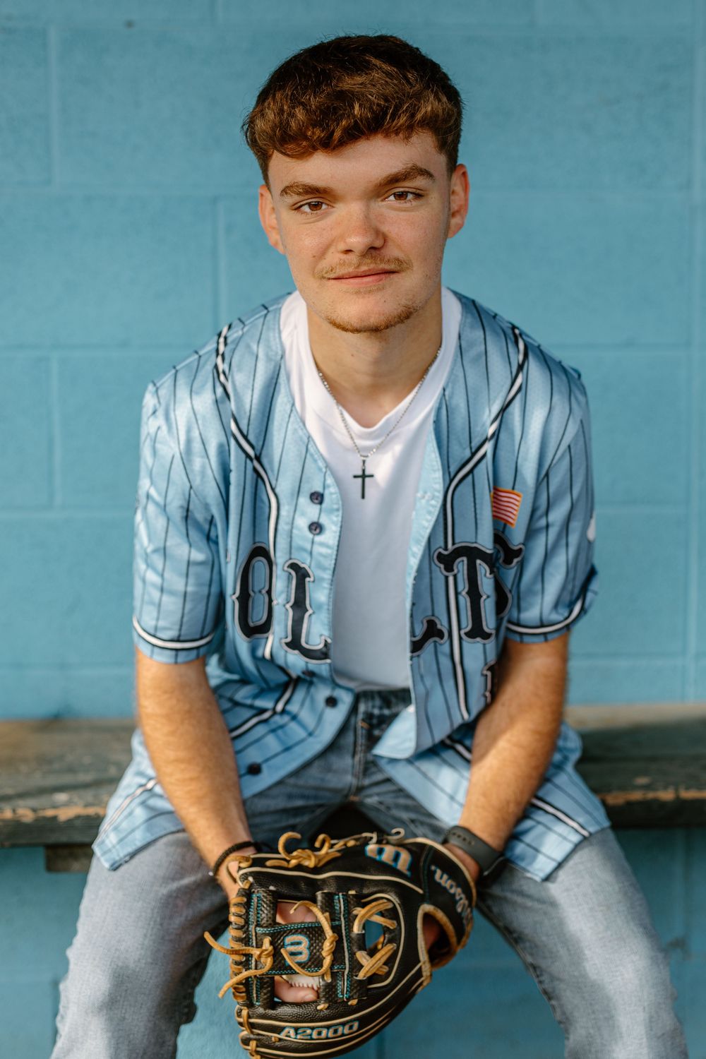 Teen baseball player in blue pinstripe jersey sitting in dugout holding A2000 glove.