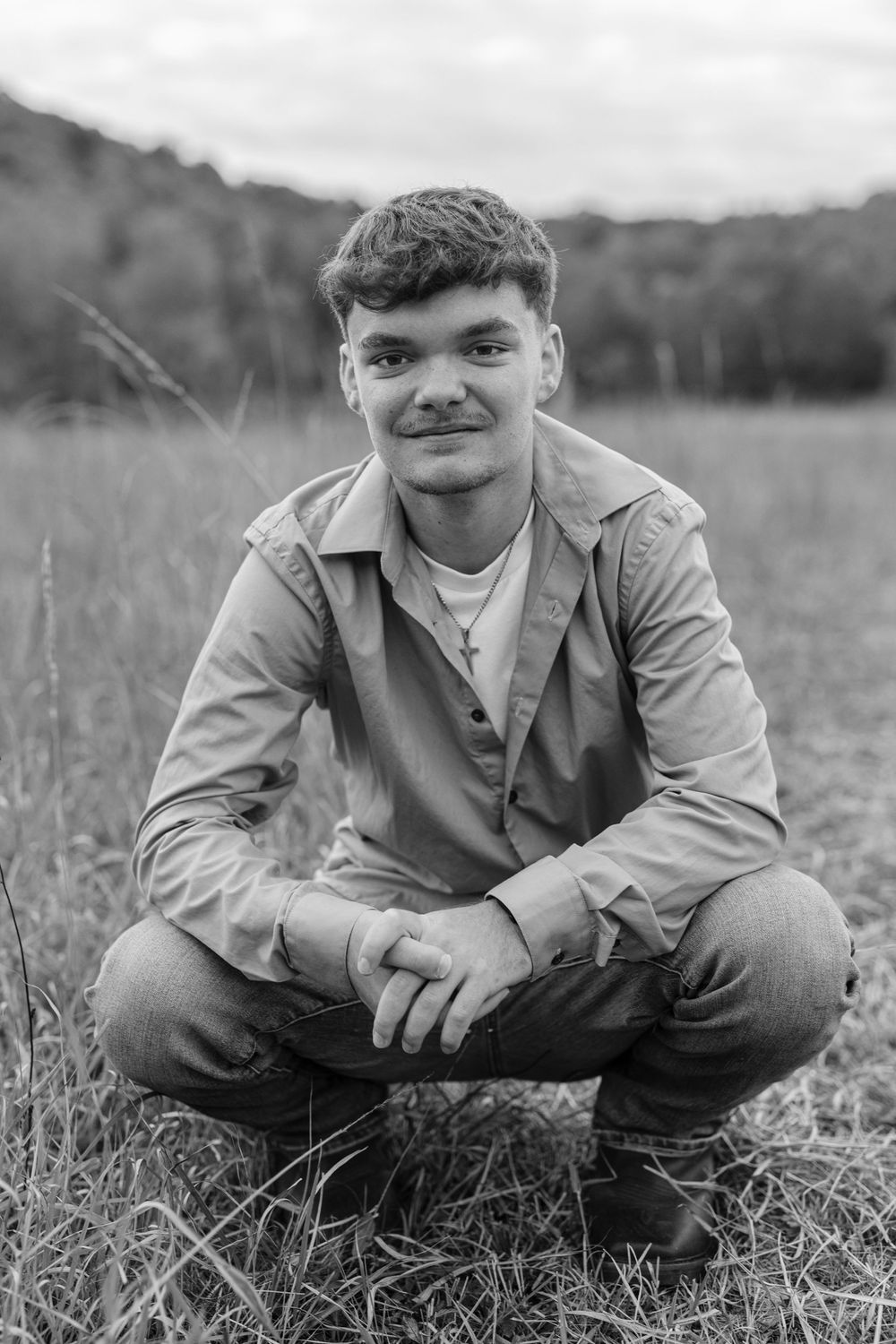 Young man in tan jacket squats in a grassy field in a black and white outdoor portrait.