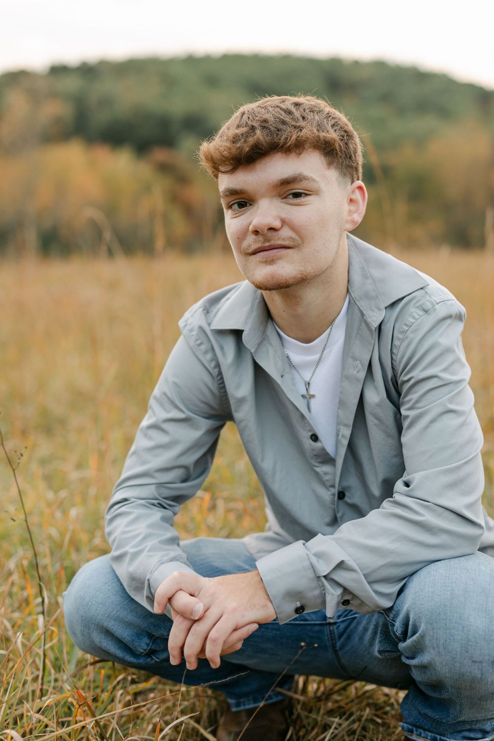 Young man in gray shirt sits in autumn field wearing cross necklace, smiling at camera.