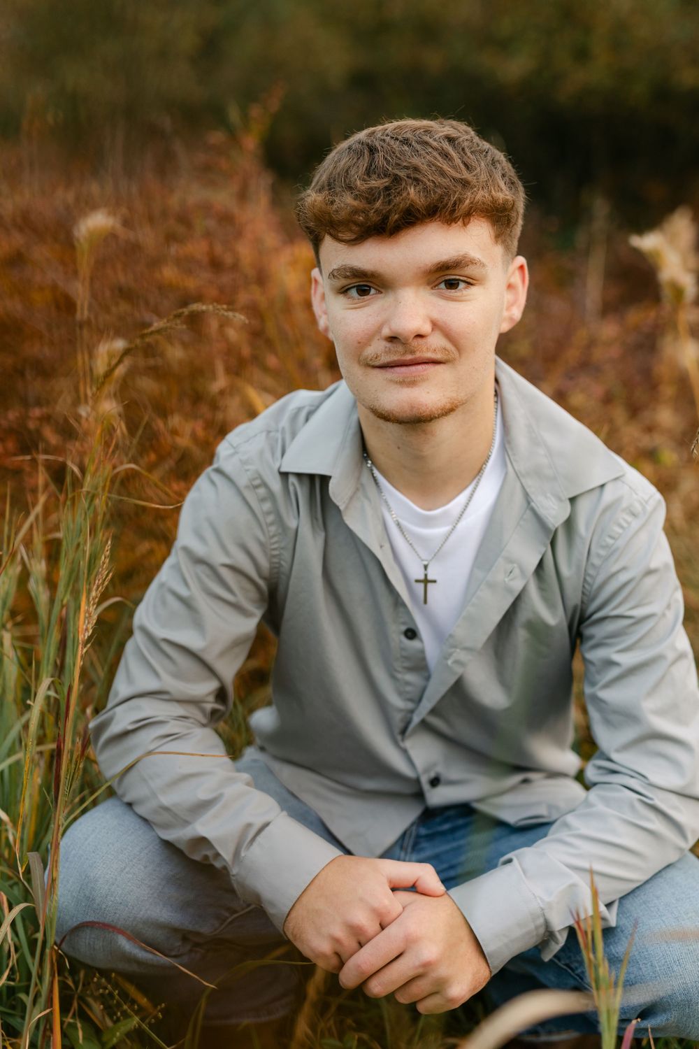 Young man in gray jacket sits in tall autumn grass wearing a cross necklace outdoors.