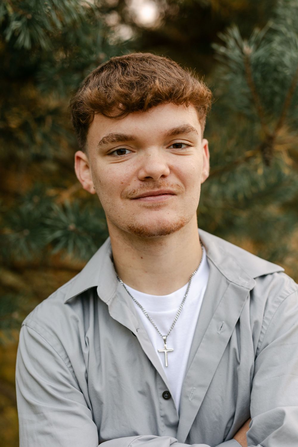 Young man in gray shirt with cross necklace smiles confidently in front of pine trees.