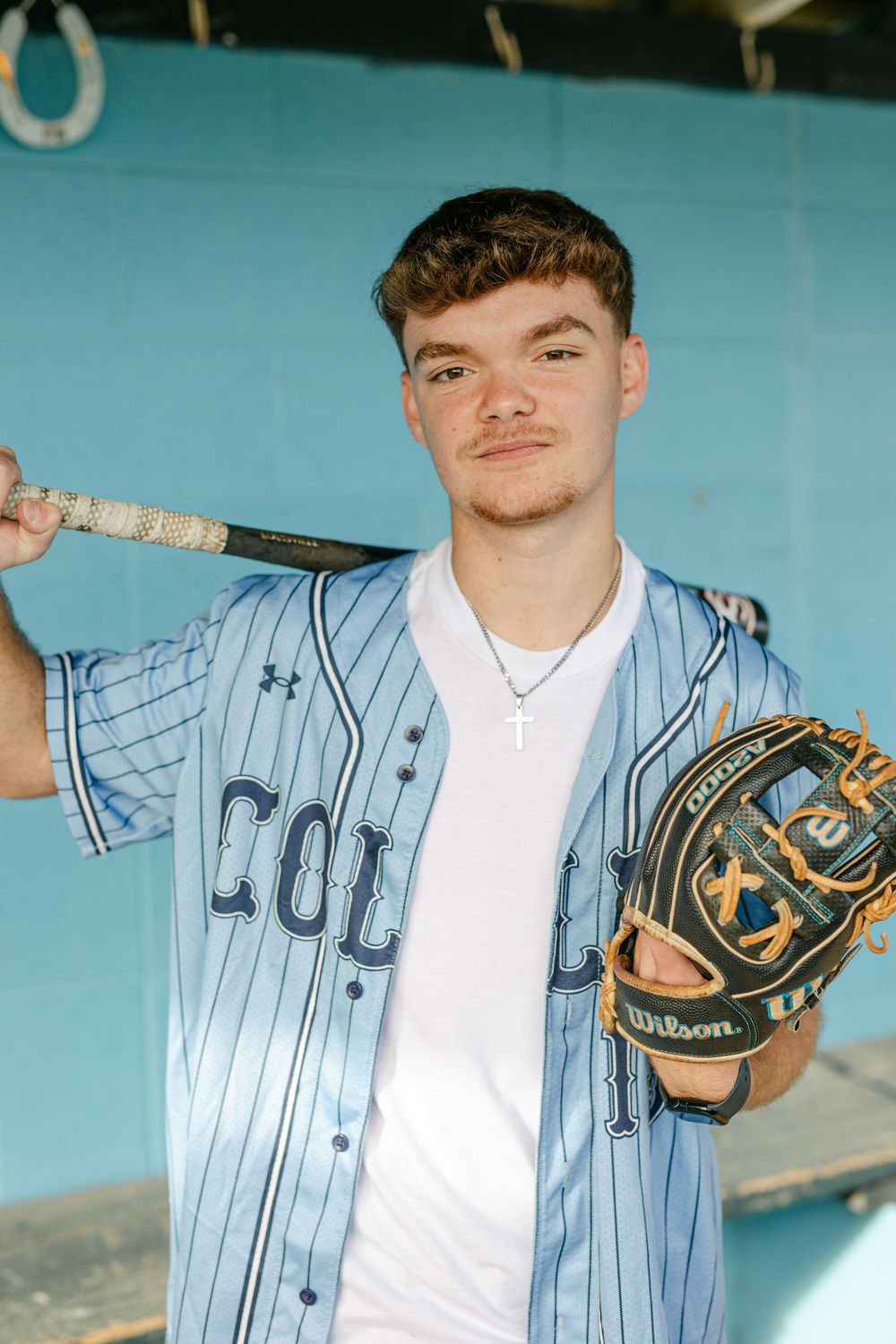 Young baseball player in blue pinstripe jersey holding bat and Wilson glove in dugout.