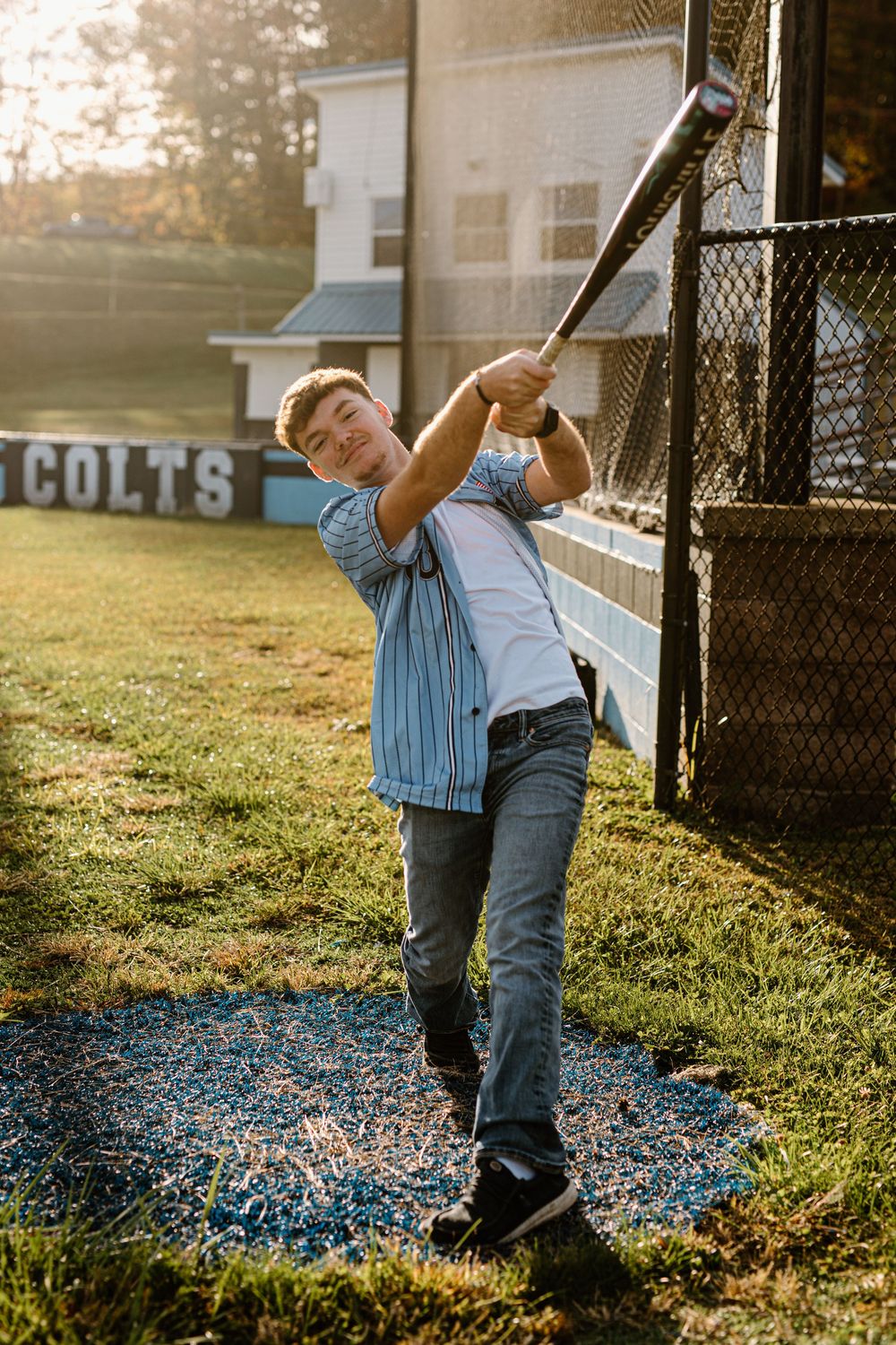 Teen baseball player in pinstripe jersey swinging bat near dugout fence in golden sunset light.