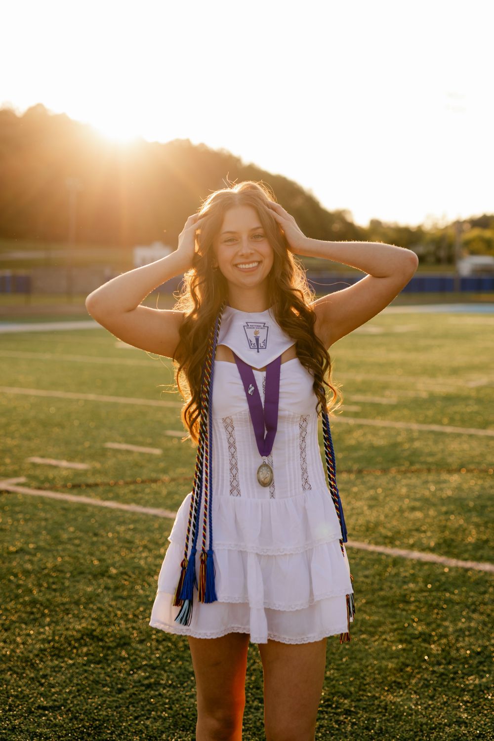 Smiling female graduate in white dress with purple medal and honor cords on sunny football field.