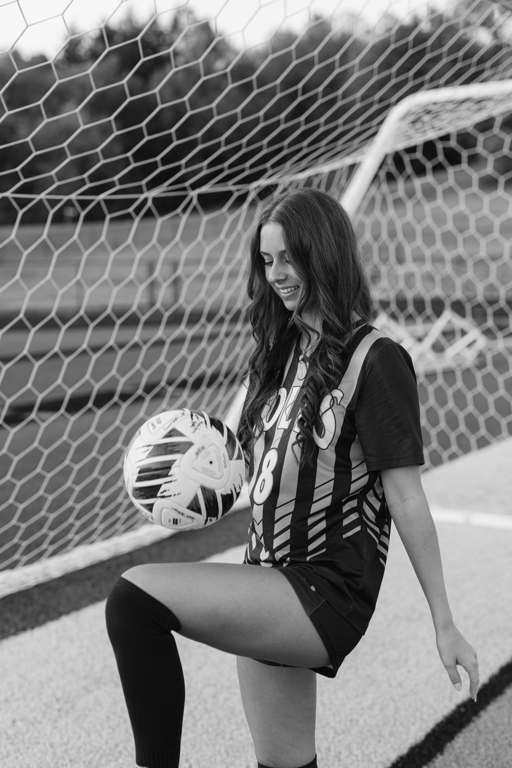 Female soccer player in Colts jersey number 8 juggling ball near goal net, black and white photo.