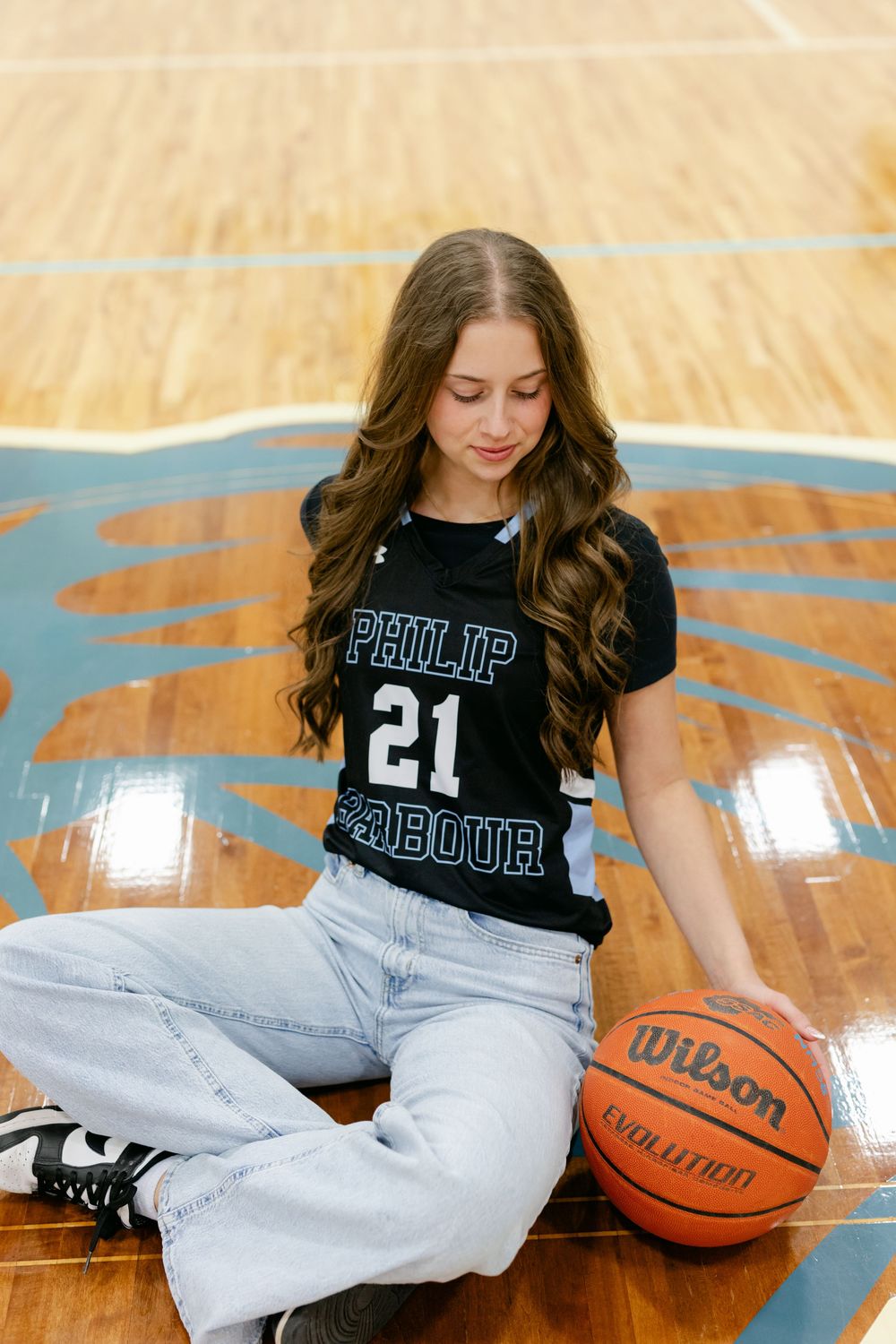 Female basketball player in Philip Barbour number 21 jersey sitting on court floor with Wilson basketball.