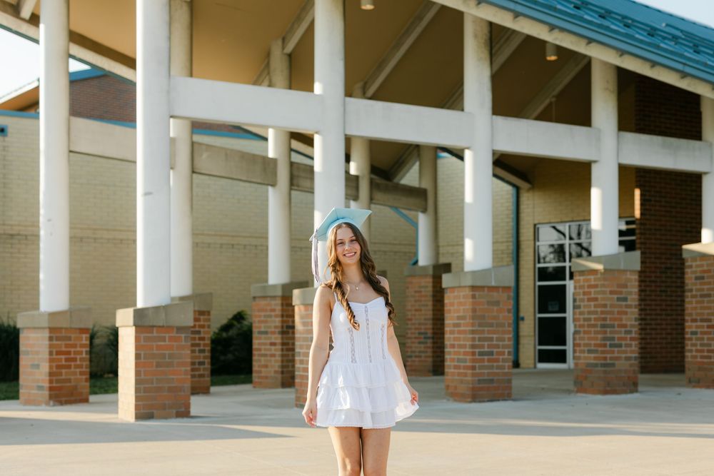 Young woman in white dress and light blue graduation cap posing outside brick school.