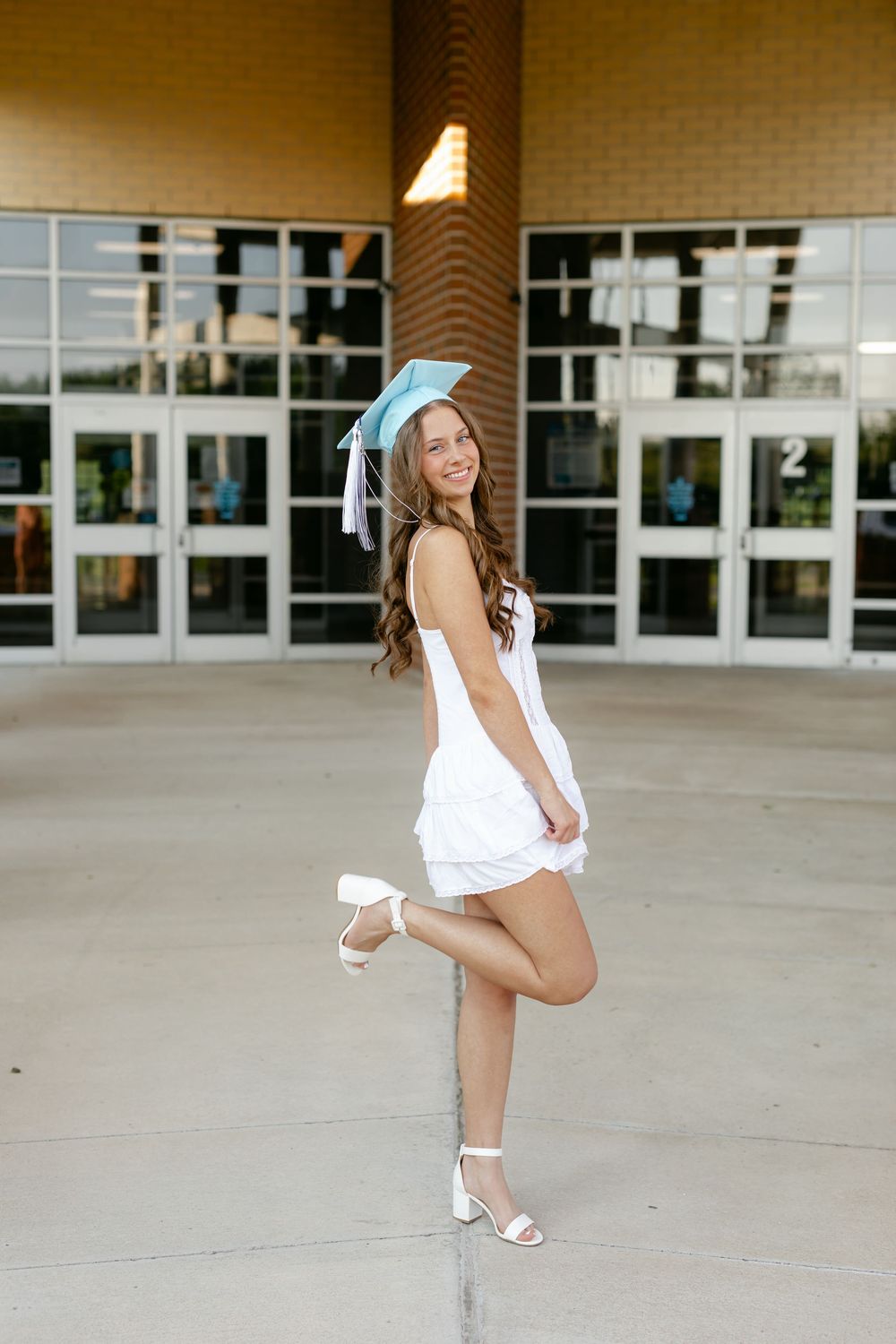 Graduate in light blue cap and white dress posing playfully outside school entrance doors.