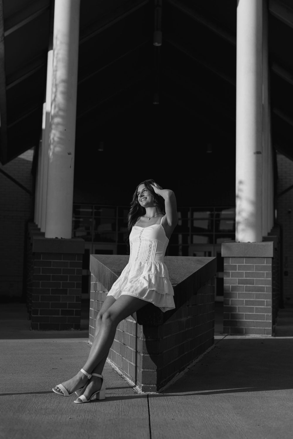 Black and white portrait of a smiling young woman in a white dress leaning against a brick pillar.