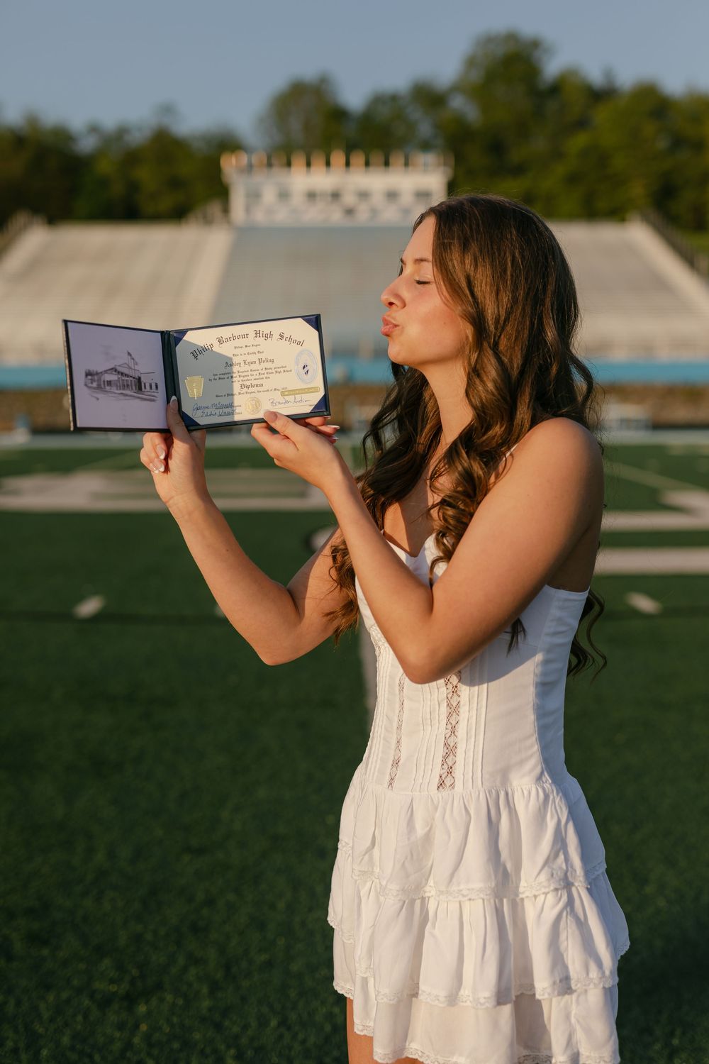 Female graduate blowing a kiss toward her open diploma on a sunny football field at golden hour.