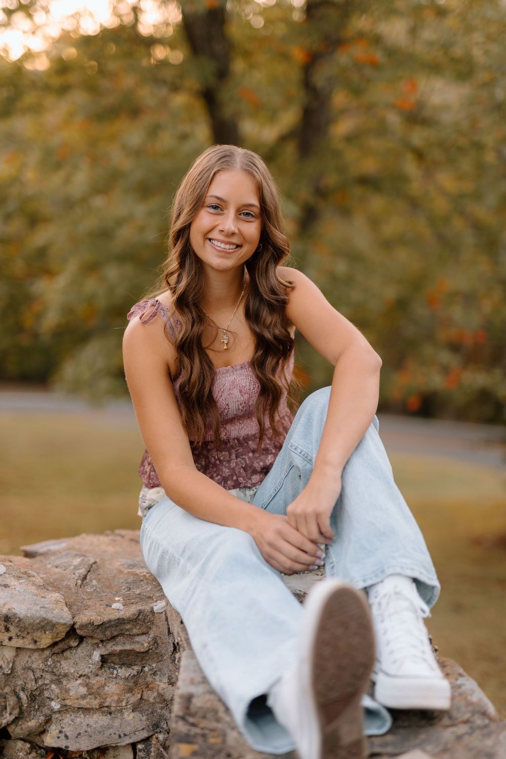 Smiling teenage girl in floral top and light jeans sitting on stone wall with autumn trees in background.