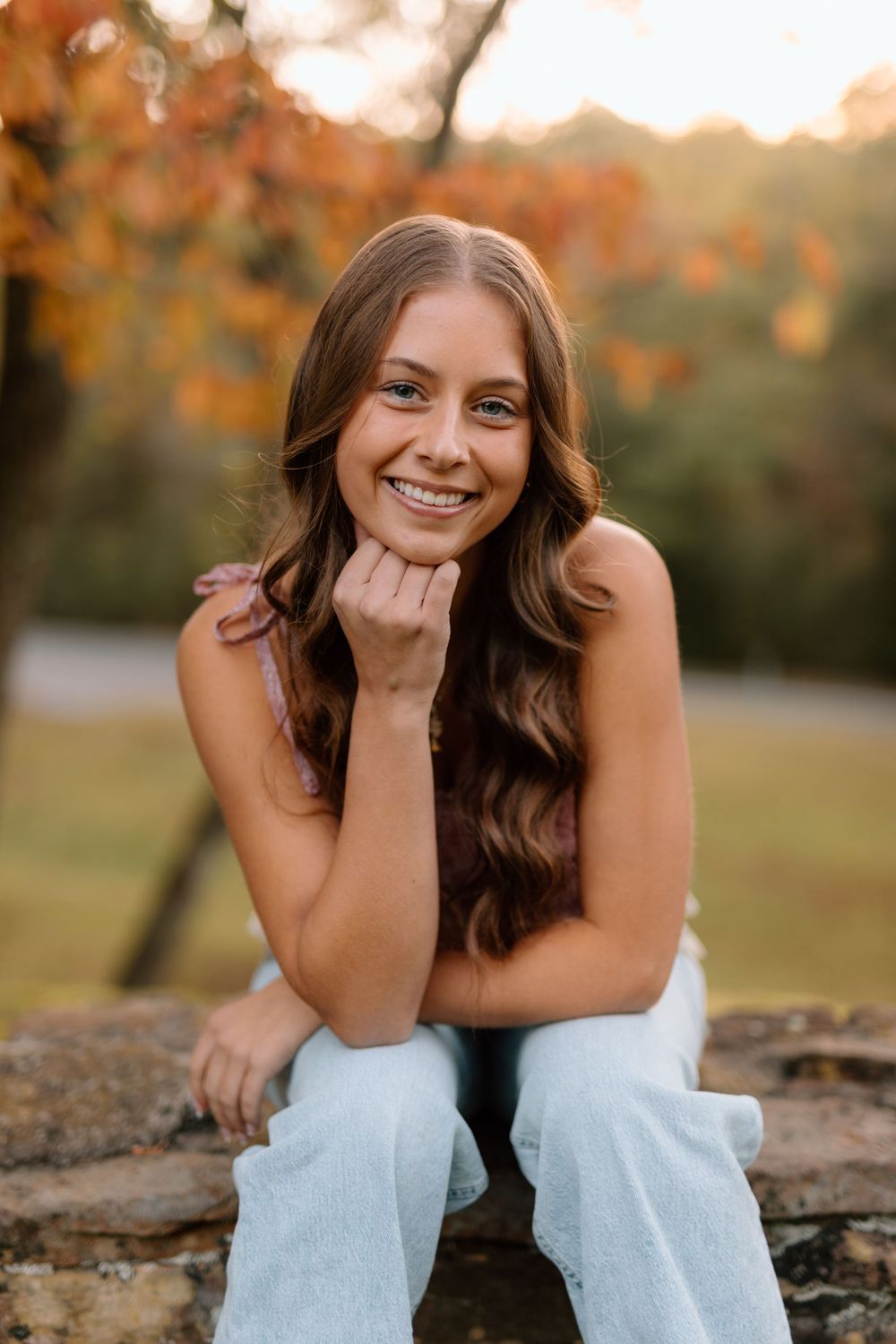 Young woman with wavy brown hair smiling warmly with chin resting on hand outdoors in fall setting.