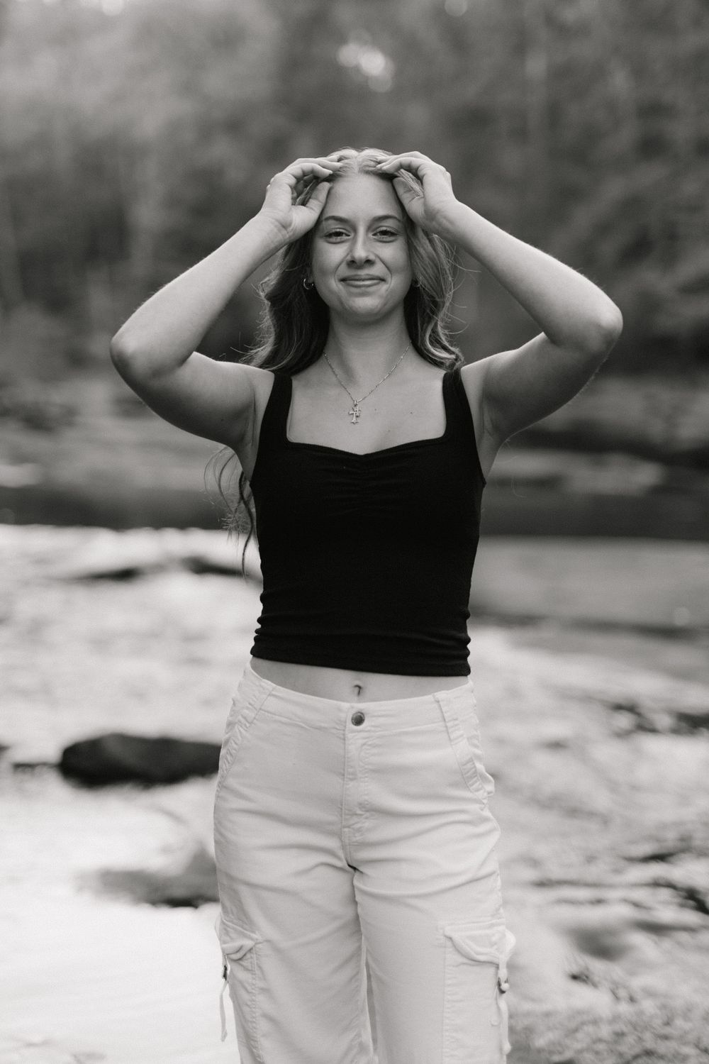 Black and white portrait of smiling girl touching her hair while standing near a flowing river outdoors.