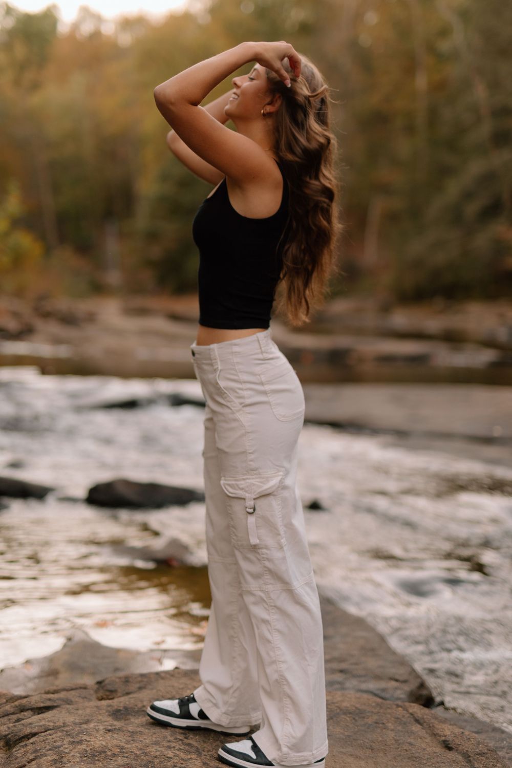Young woman in black crop top and white cargo pants posing by rocky riverside in autumn forest.
