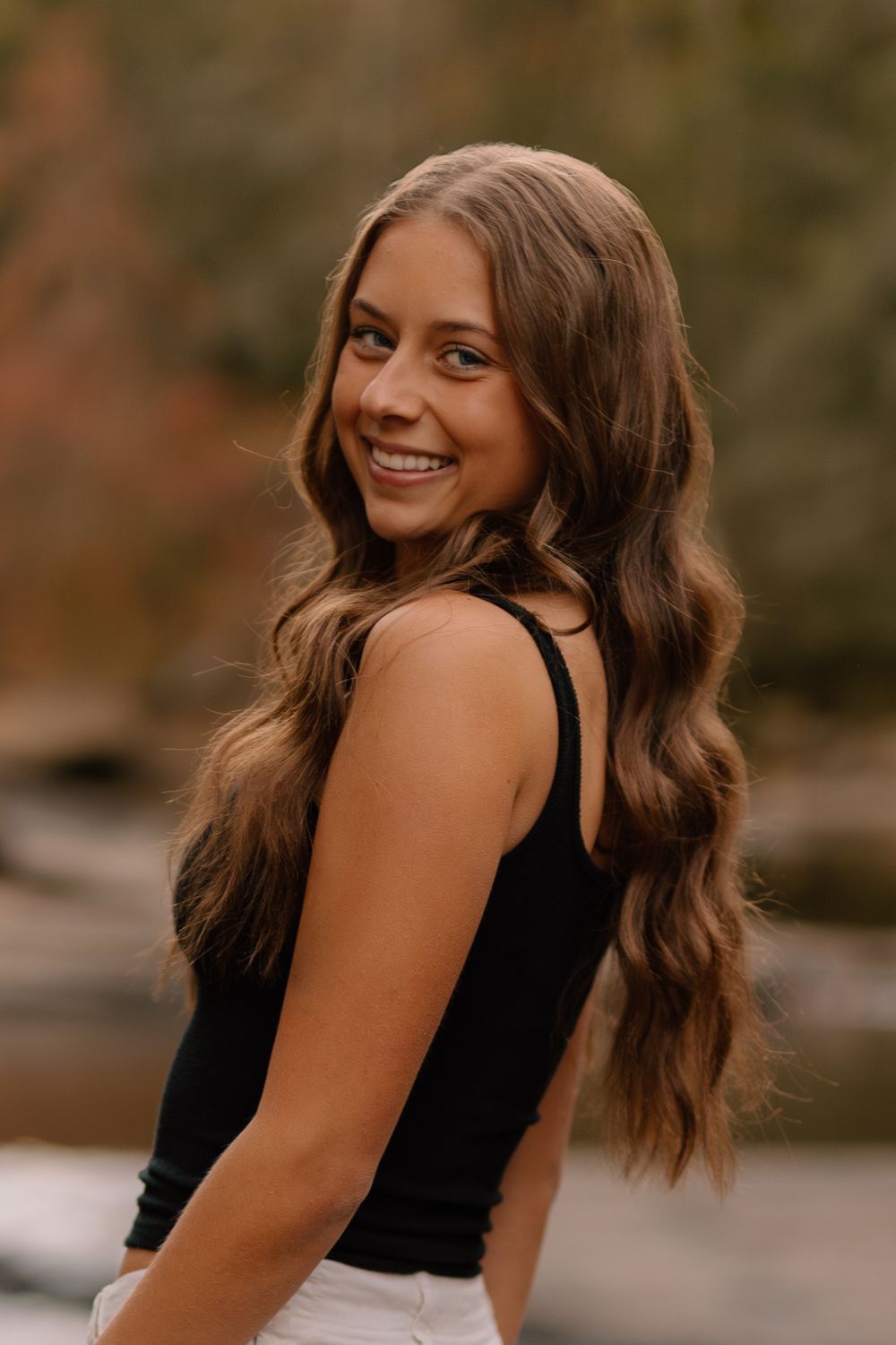 Smiling teenage girl with long wavy brown hair in black tank top glancing back near a river.