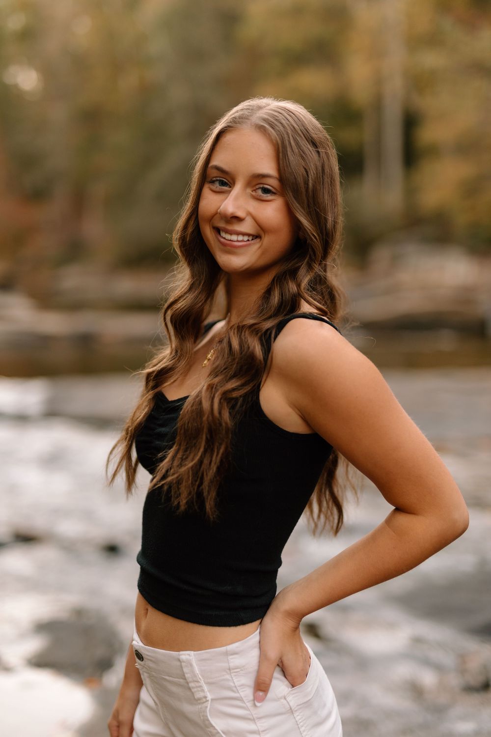 Smiling young woman in black crop top and white pants posing by a rocky river in fall setting.