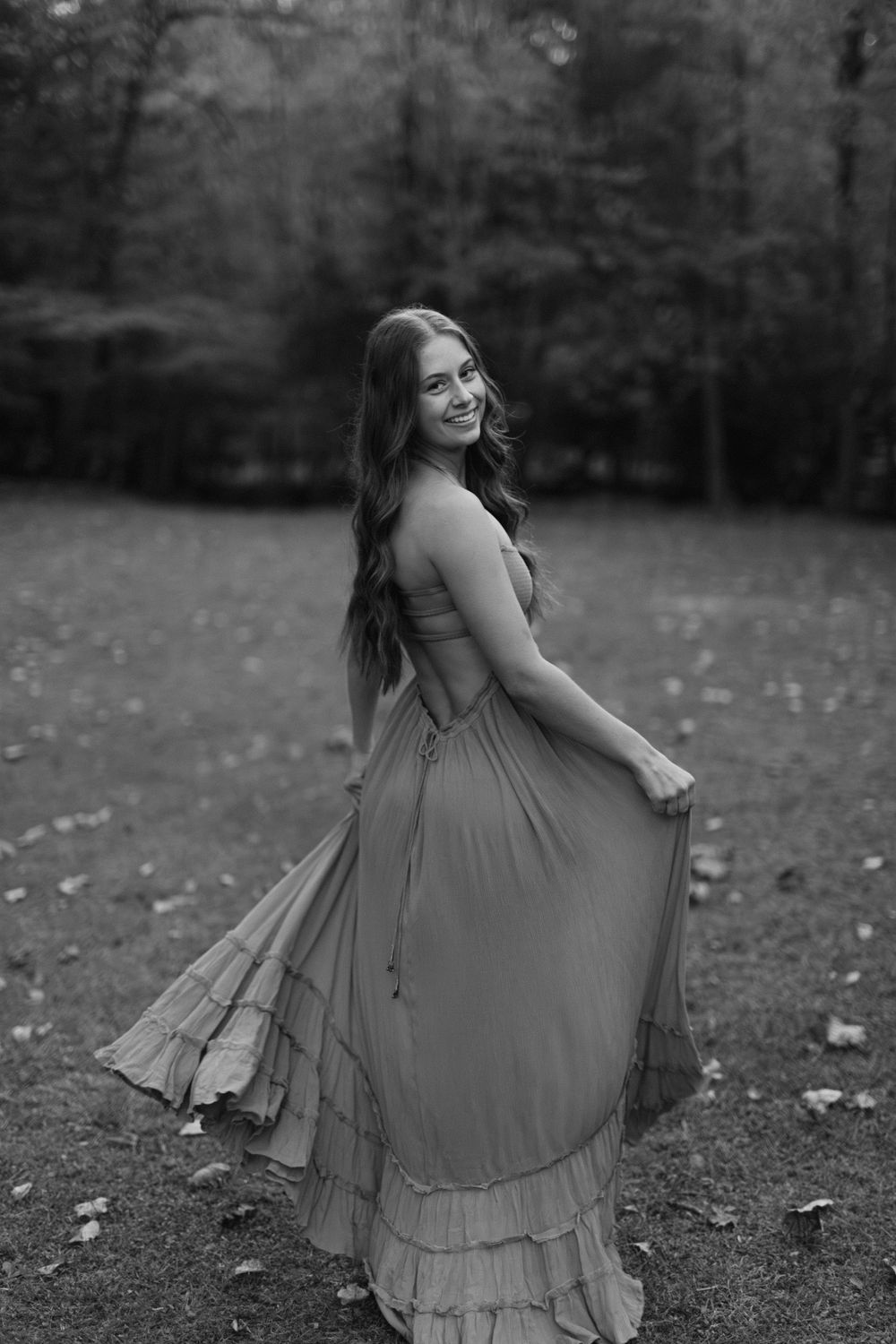 Black and white photo of young woman in flowing maxi dress spinning in a wooded outdoor setting.