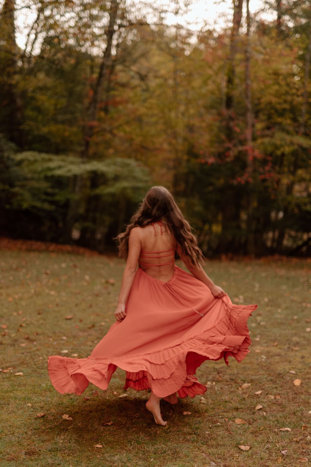 Young woman spinning in a coral ruffled maxi dress outdoors in an autumn forest setting.