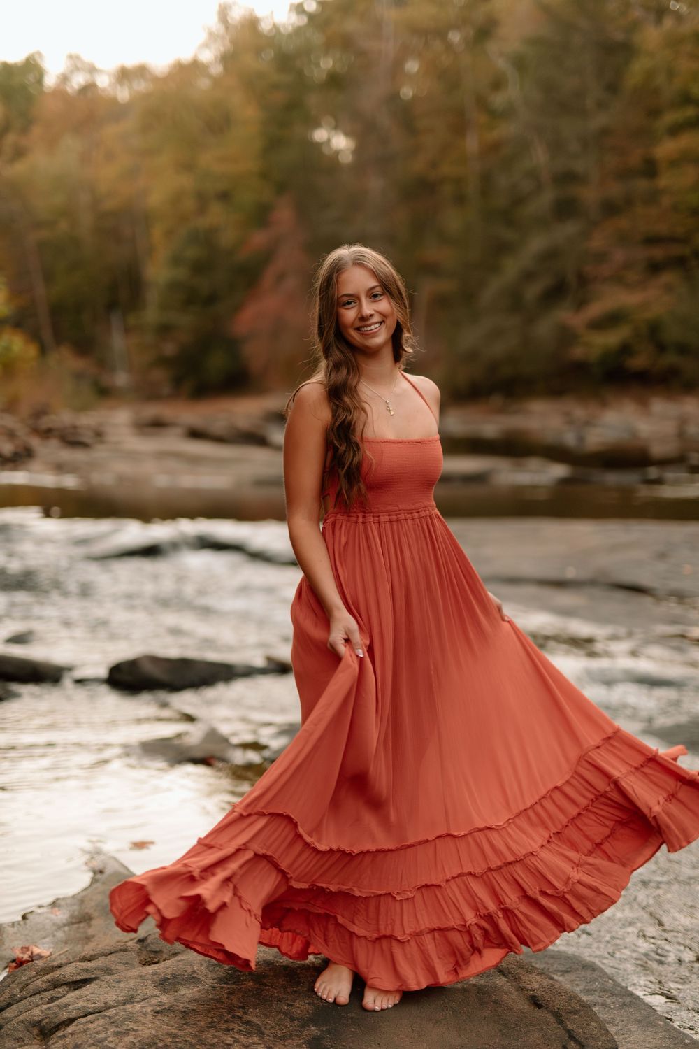 Young woman in a flowing coral ruffled maxi dress posing on rocks near an autumn river.