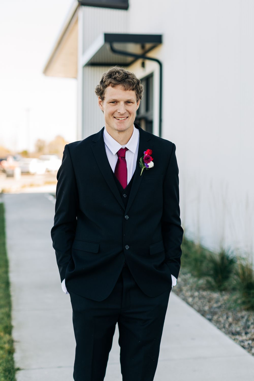 Smiling groom in black three-piece suit with burgundy tie and floral boutonniere standing outside wedding venue.
