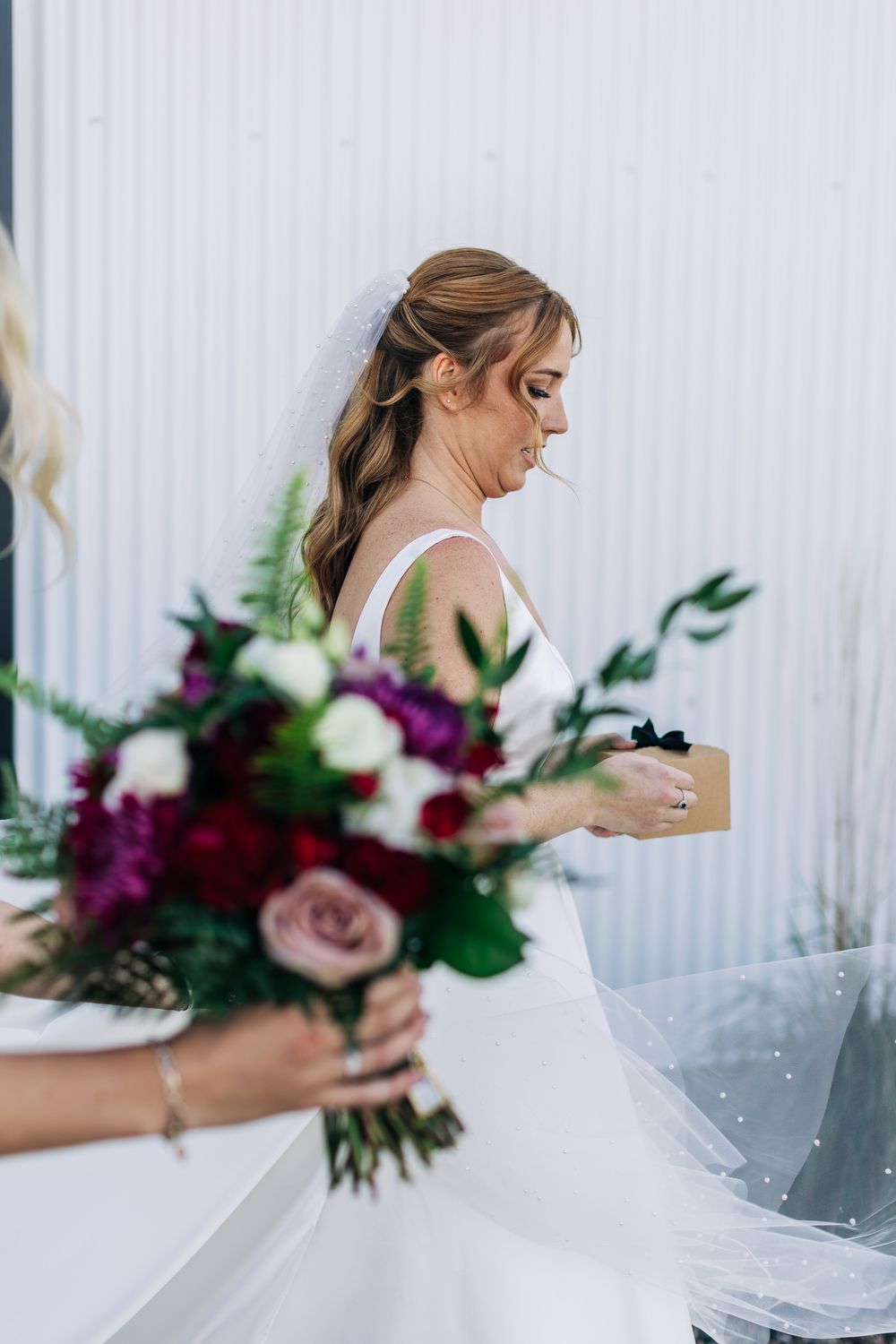 Bride in white gown holds a gift box while a colorful bouquet of red and pink roses is held in the foreground.
