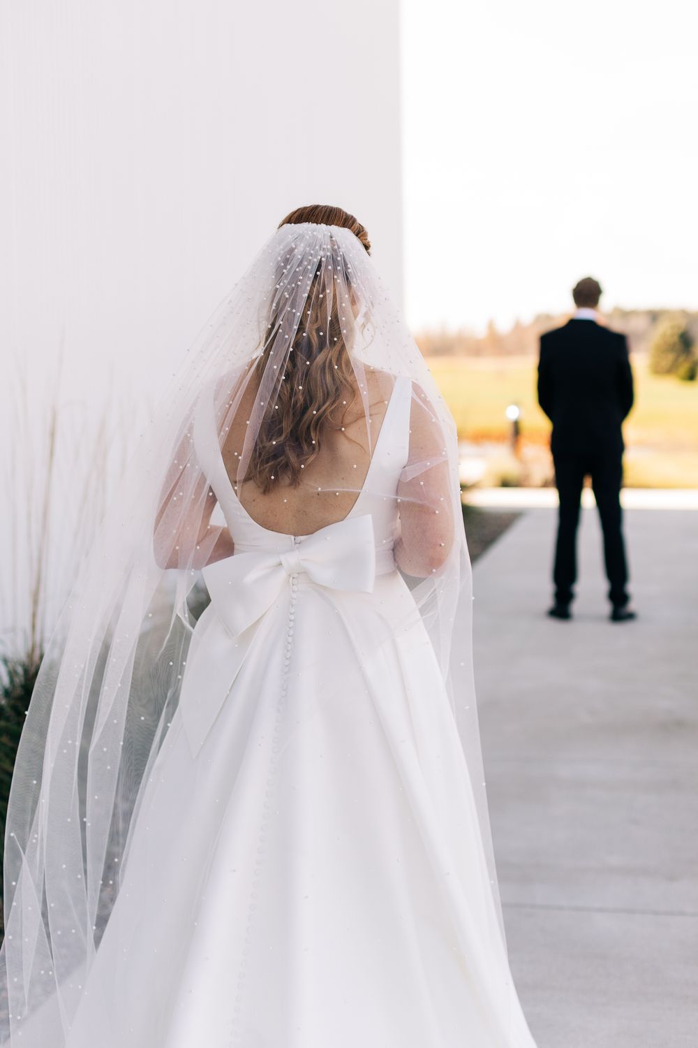 Bride in white gown with pearl veil and bow detail approaches groom for first look.