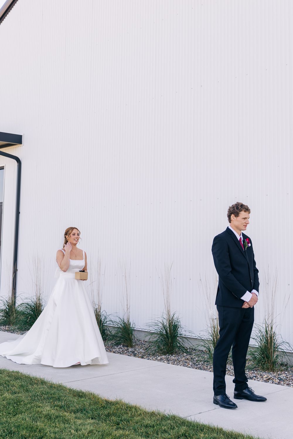 Bride in white gown approaches groom in black suit during outdoor first look at wedding venue.
