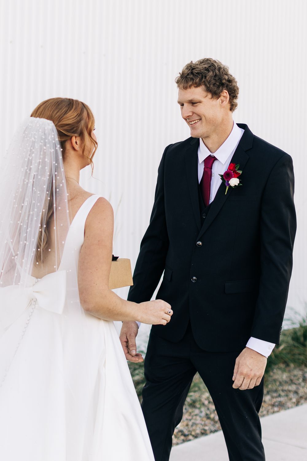 Groom in black suit with red tie smiles at bride during wedding first look moment.