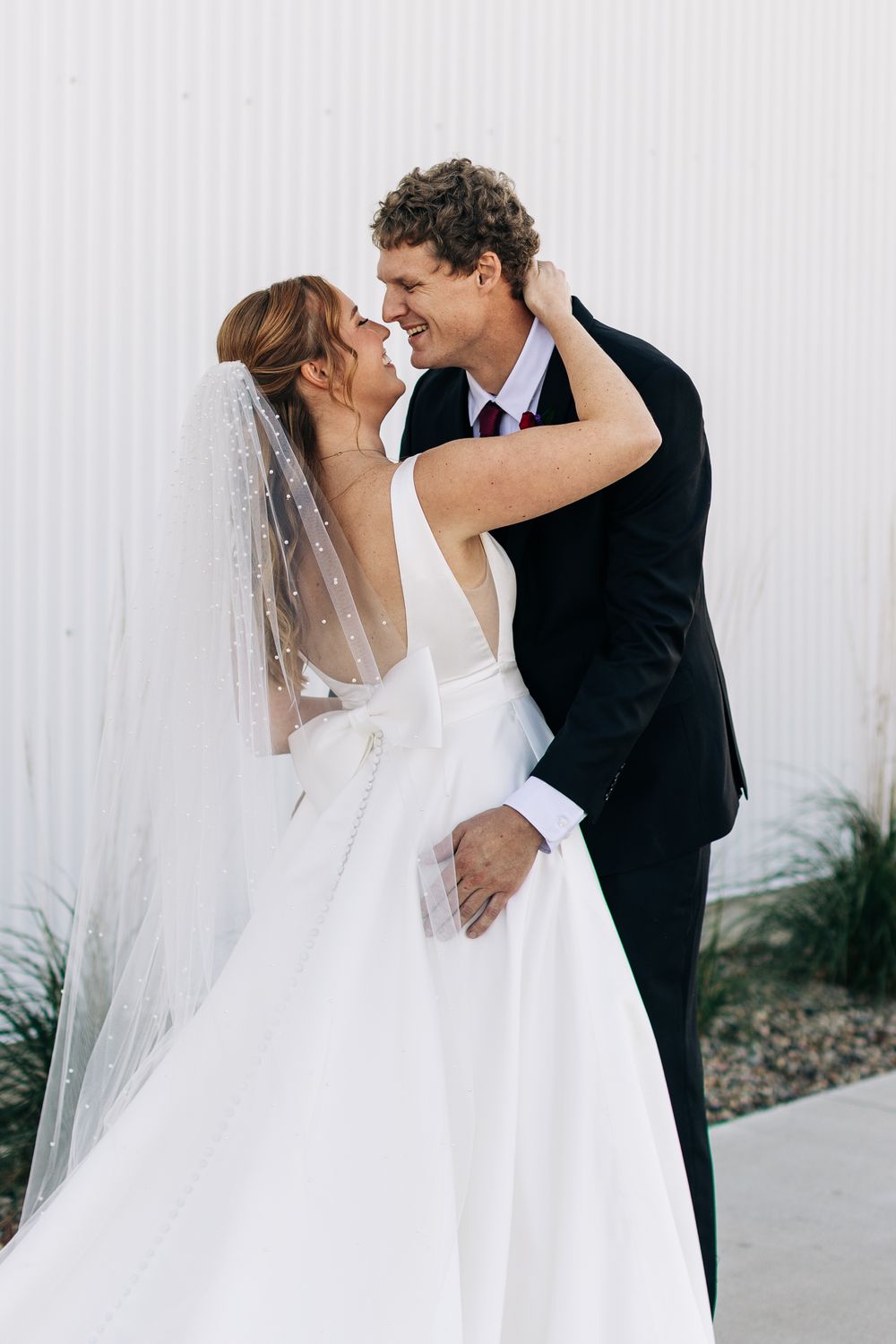 Bride and groom share a romantic moment, nearly kissing, as she wears a white backless gown with veil outdoors.