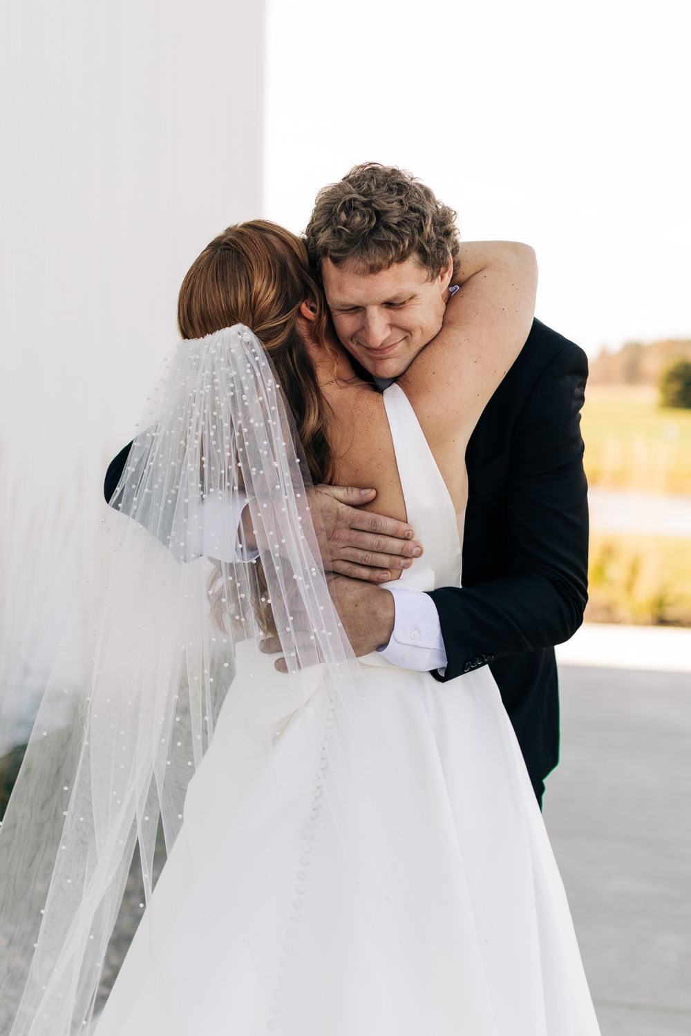 Bride and groom share emotional first look embrace outside white wedding venue.