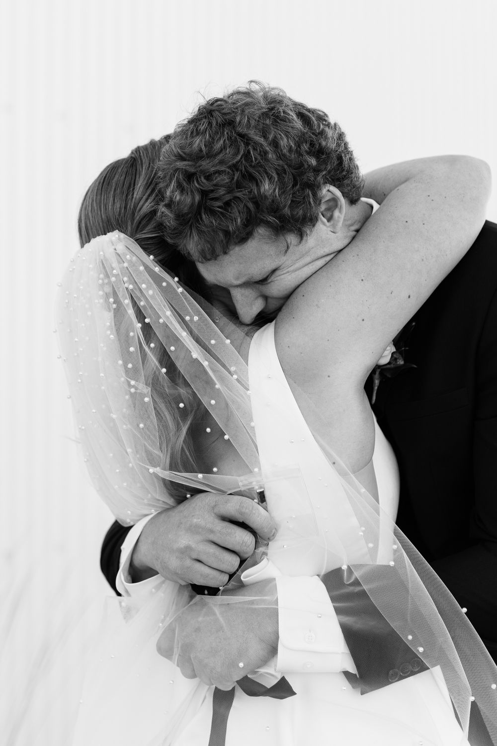 Bride and groom share an intimate embrace in a black and white wedding photo, her veil draped elegantly.